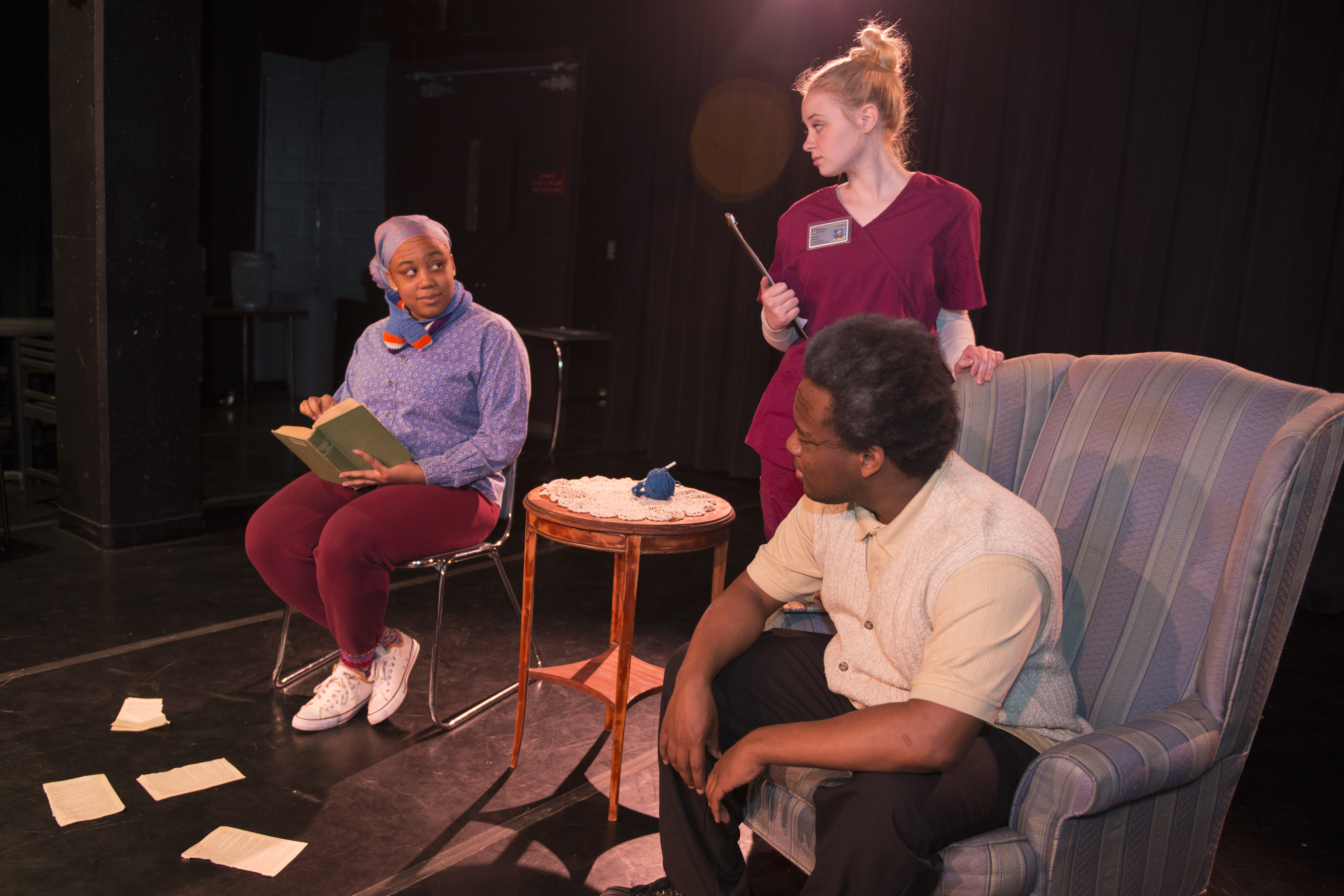 Three actors, two are sitting in a chair next to a side table