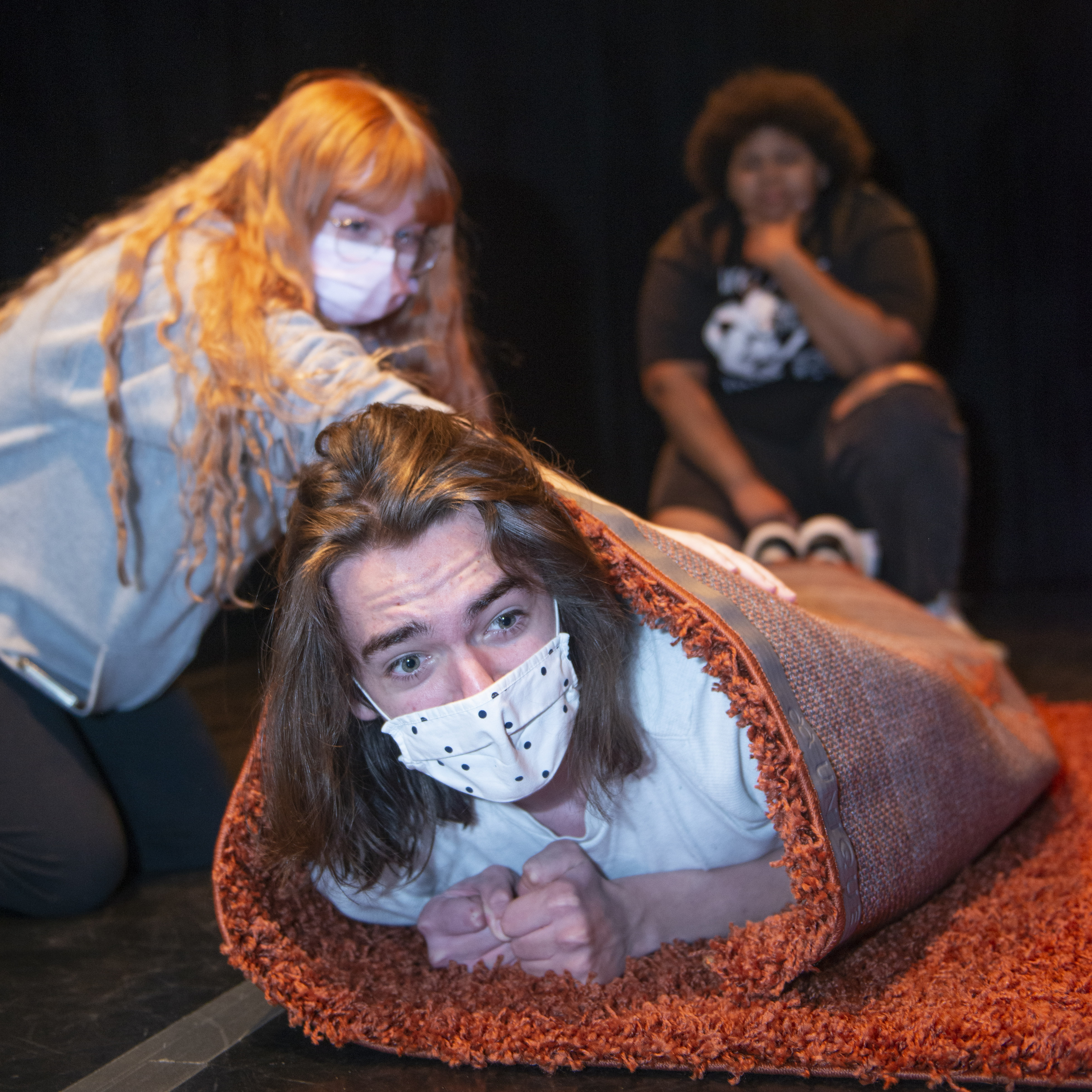 Three theater students one of them laying down wrapped up in a carpet