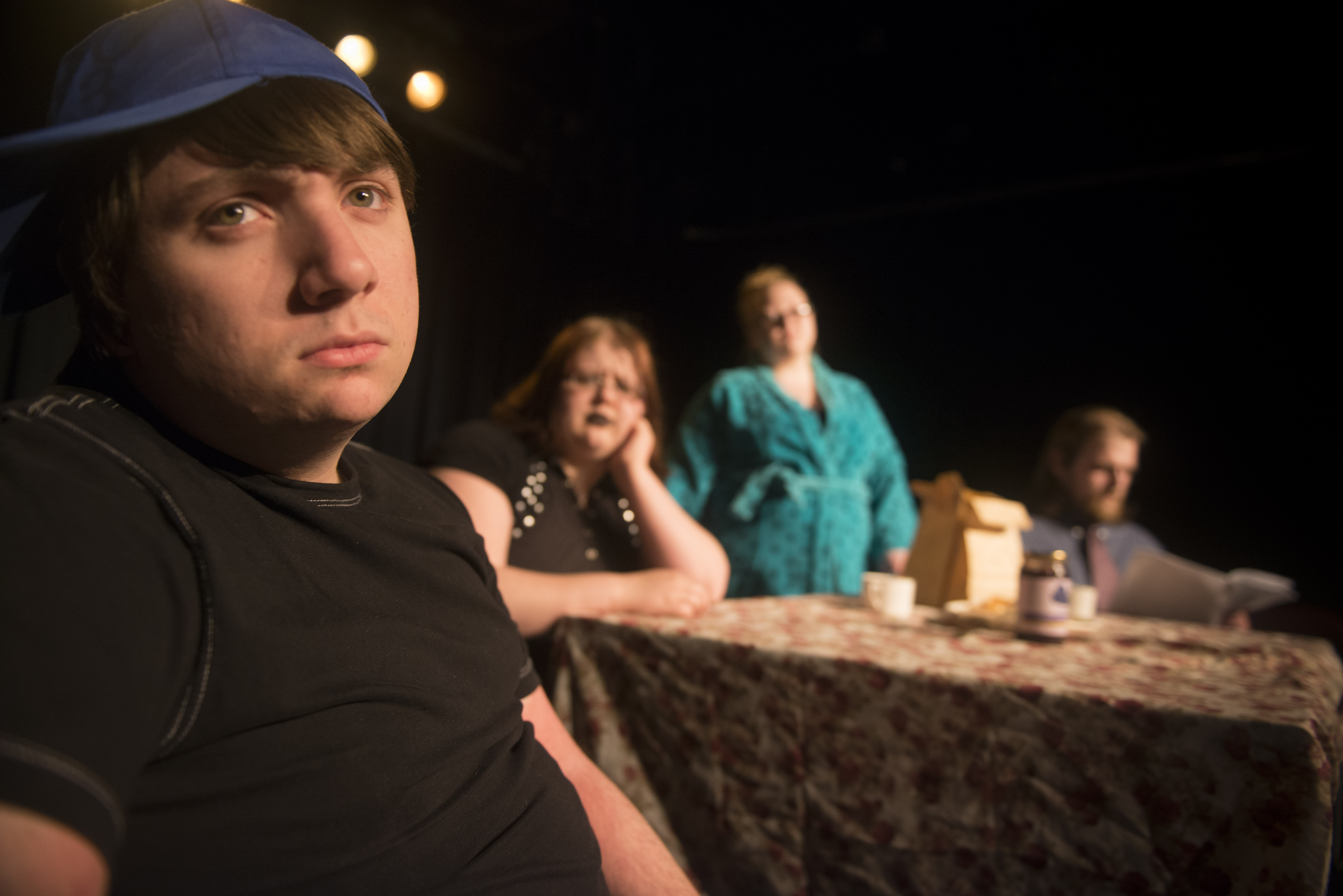 Four actors staring at the camera. Closest one is wearing a black shite with a blue baseball hat