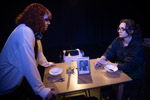 Two theater students at a table. One is sitting down the other is standing over the table