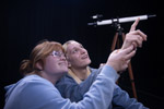 Two theater students pointing up at the sky with a telescope behind them