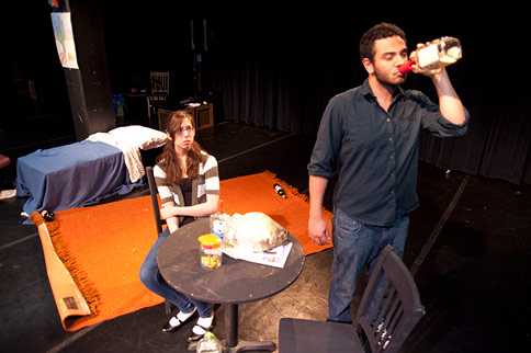 Female actress sitting at at a table staring at male actor drinking out of a bottle
