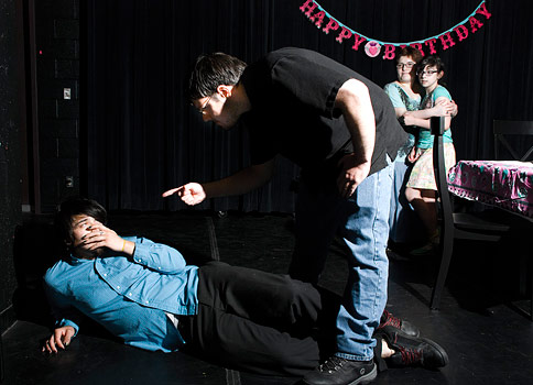 Actor wearing blue shirt laying on the ground while actor wearing black shirt standing over him pointing