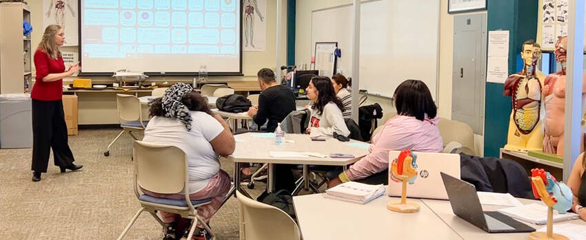 Female speaker talking to students at the Natural Science Education Center classroom, with tables, chairs, and human anatomy figure and illustrations