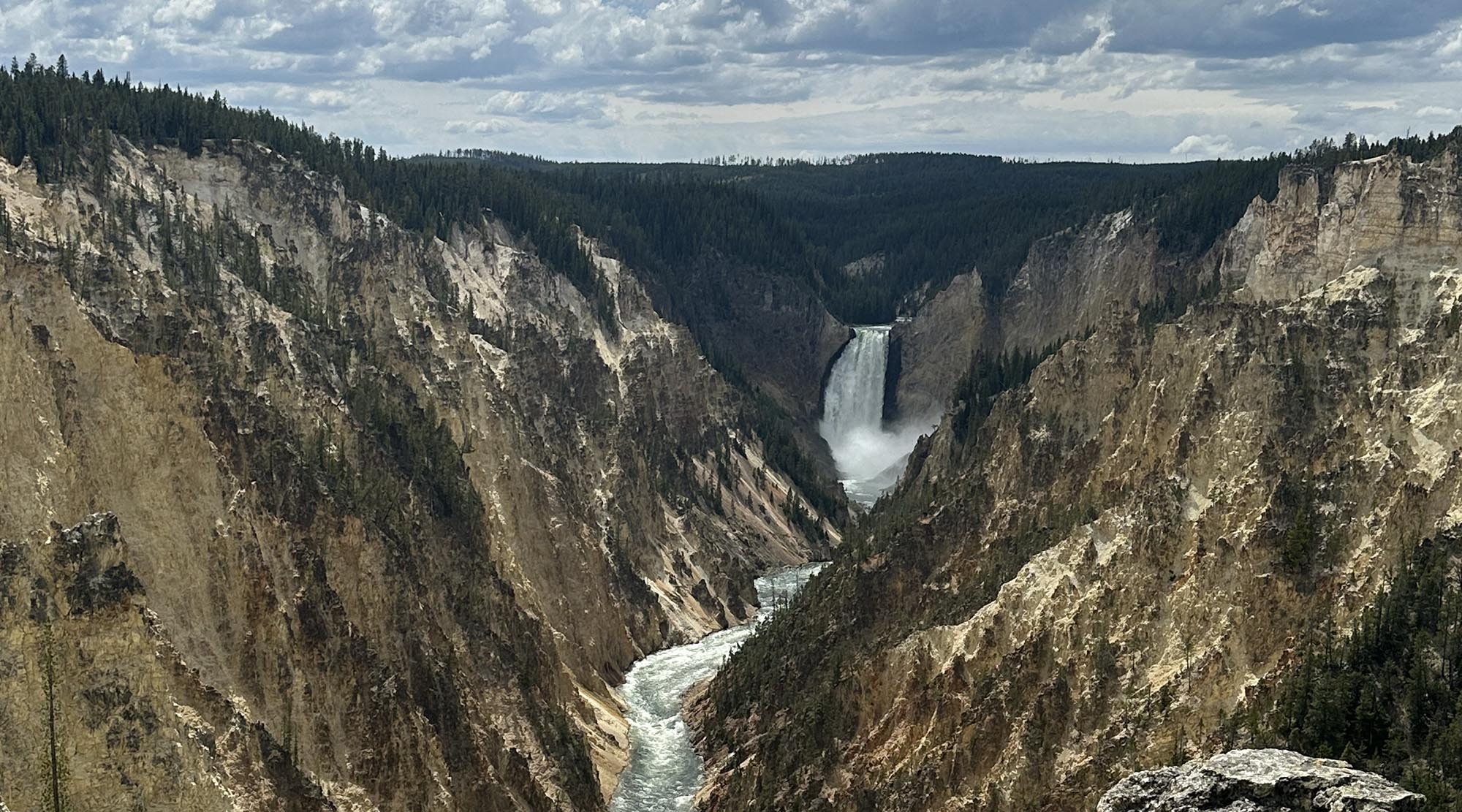 A wide scenic view of a deep, rugged canyon with steep, multicolored rock walls and dense evergreen forests. A tall waterfall plunges into the canyon near the center, with a winding whitewater river flowing through the gorge below.