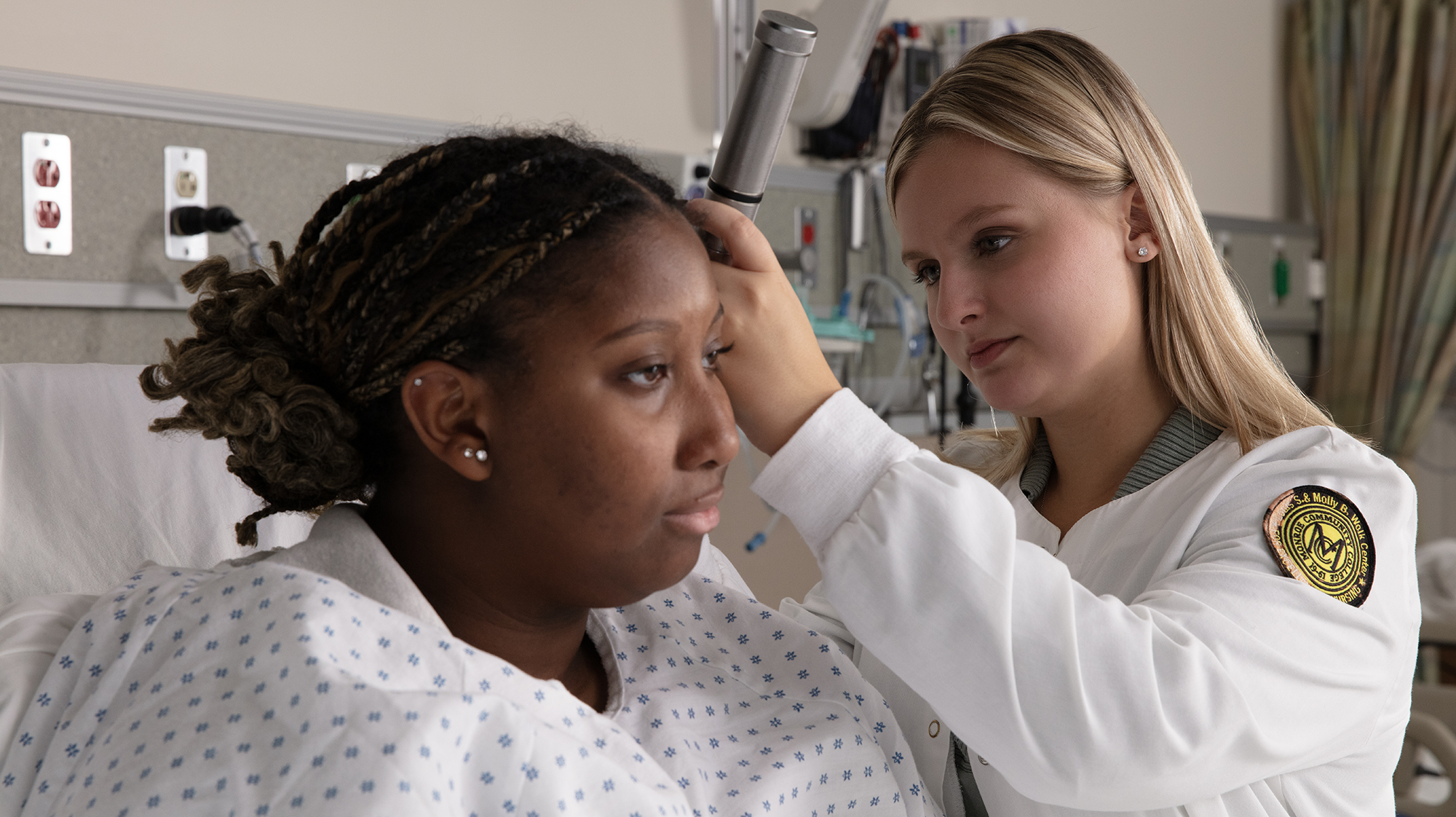 A health studies student in a white clinical coat examines a seated patient with an otoscope in a simulated hospital room during a hands-on clinical training session.