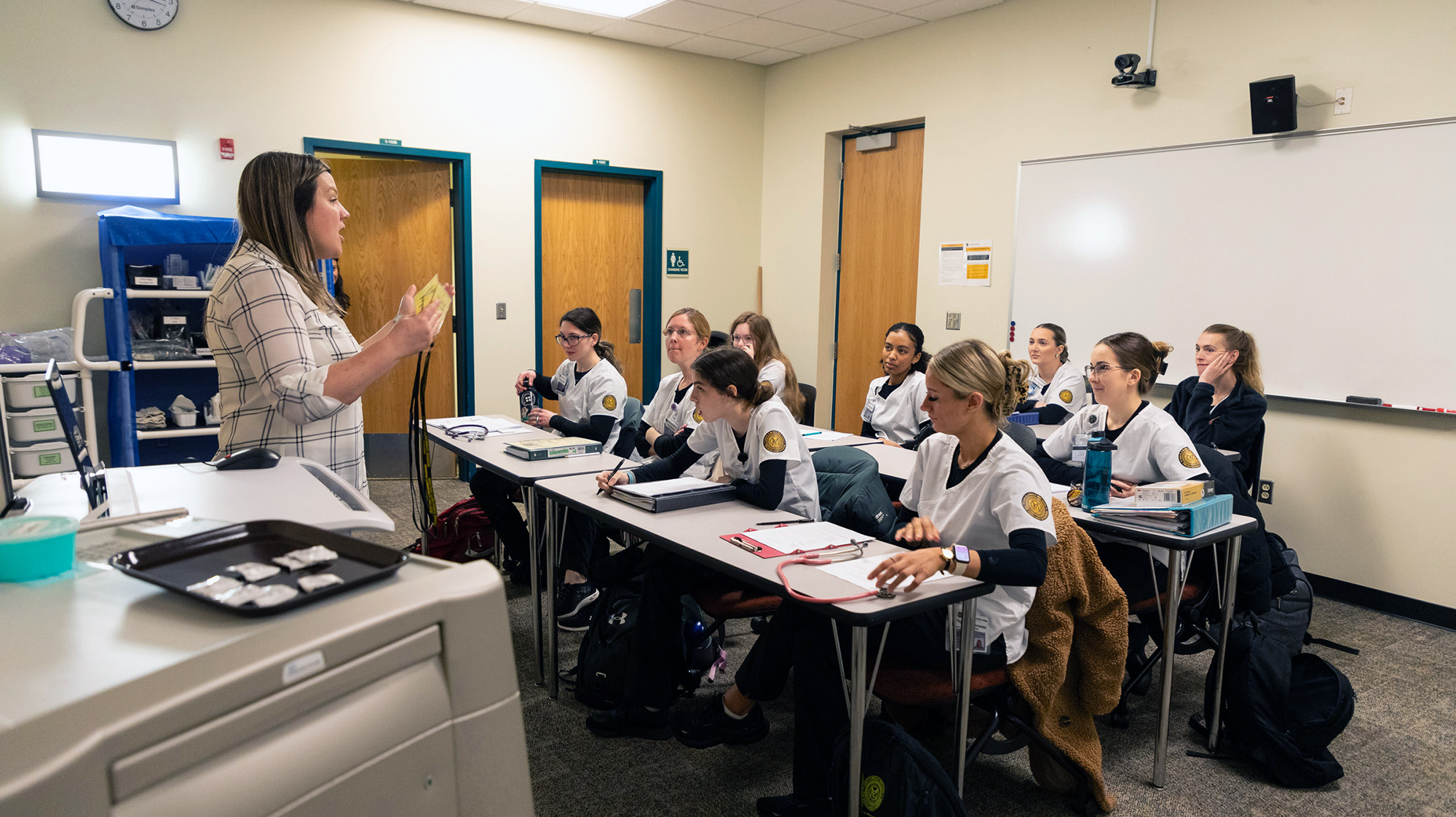 Instructor teaches a classroom of healthcare students in scrubs during a hands-on lesson.