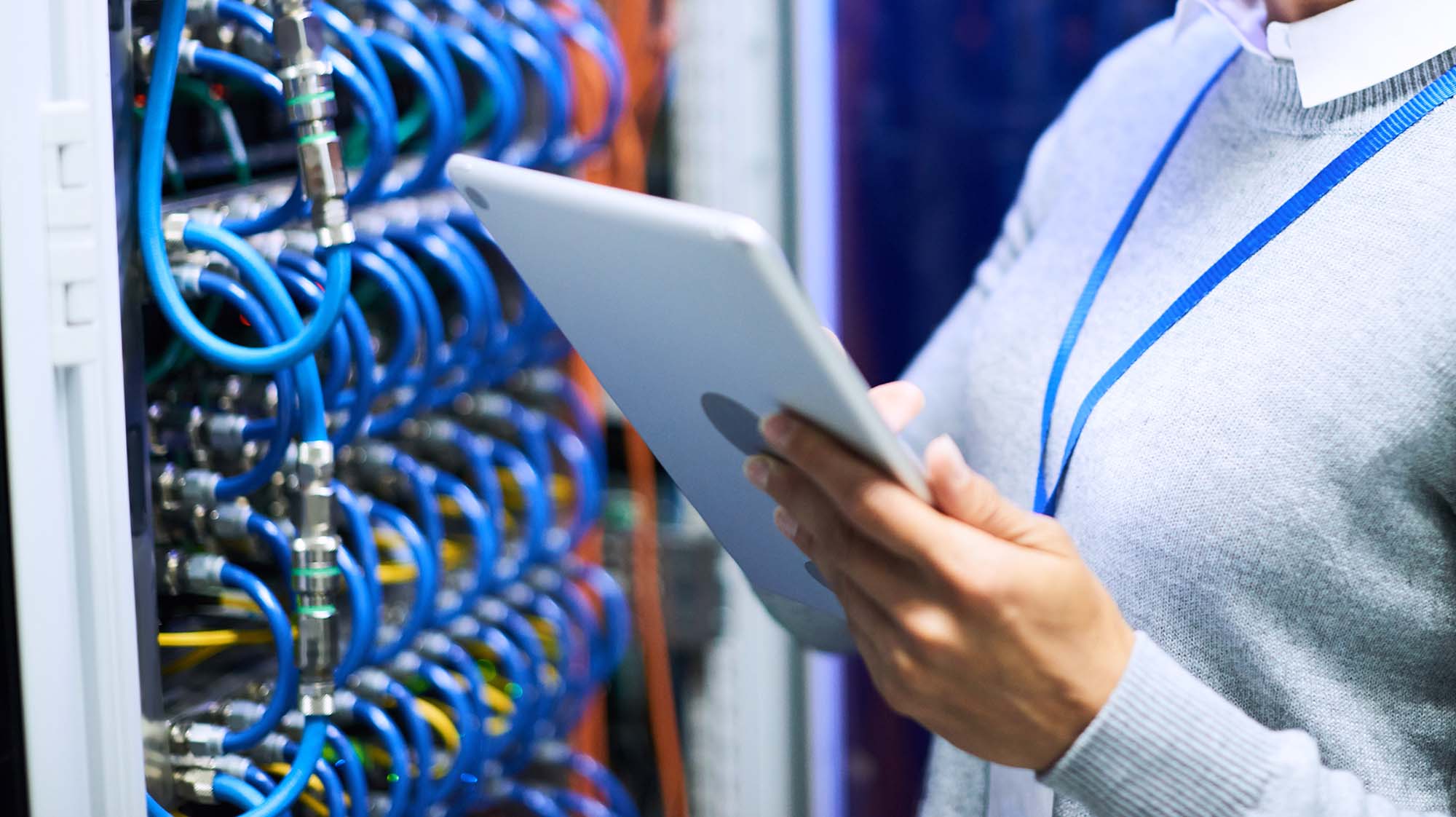 Person uses a tablet to monitor and manage network equipment in a server room with connected network cables.