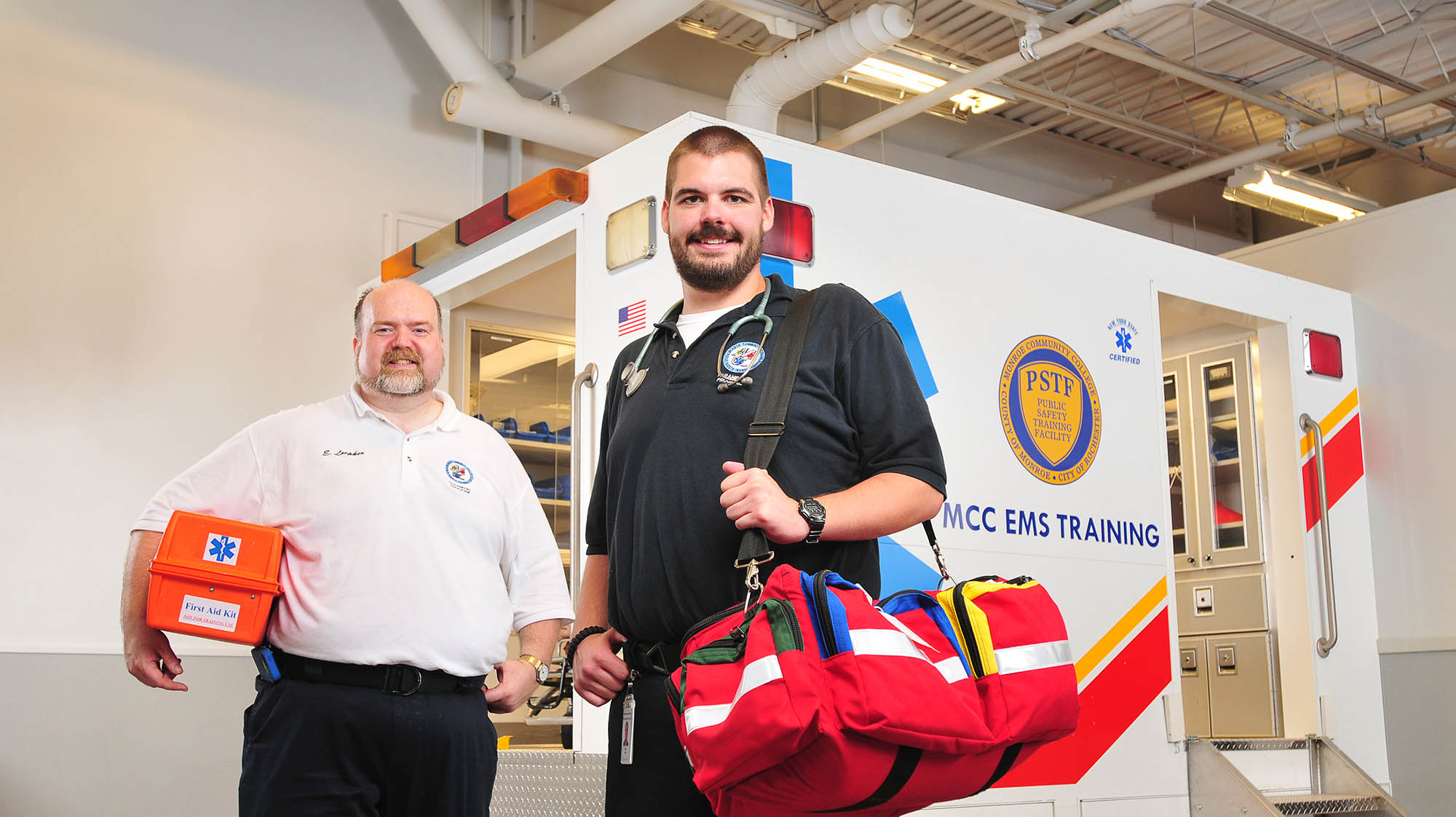 Two EMS professionals pose in front of an ambulance training simulator at Monroe Community College. One holds a first aid kit, while the other wears EMS gear and carries a red medical response bag.