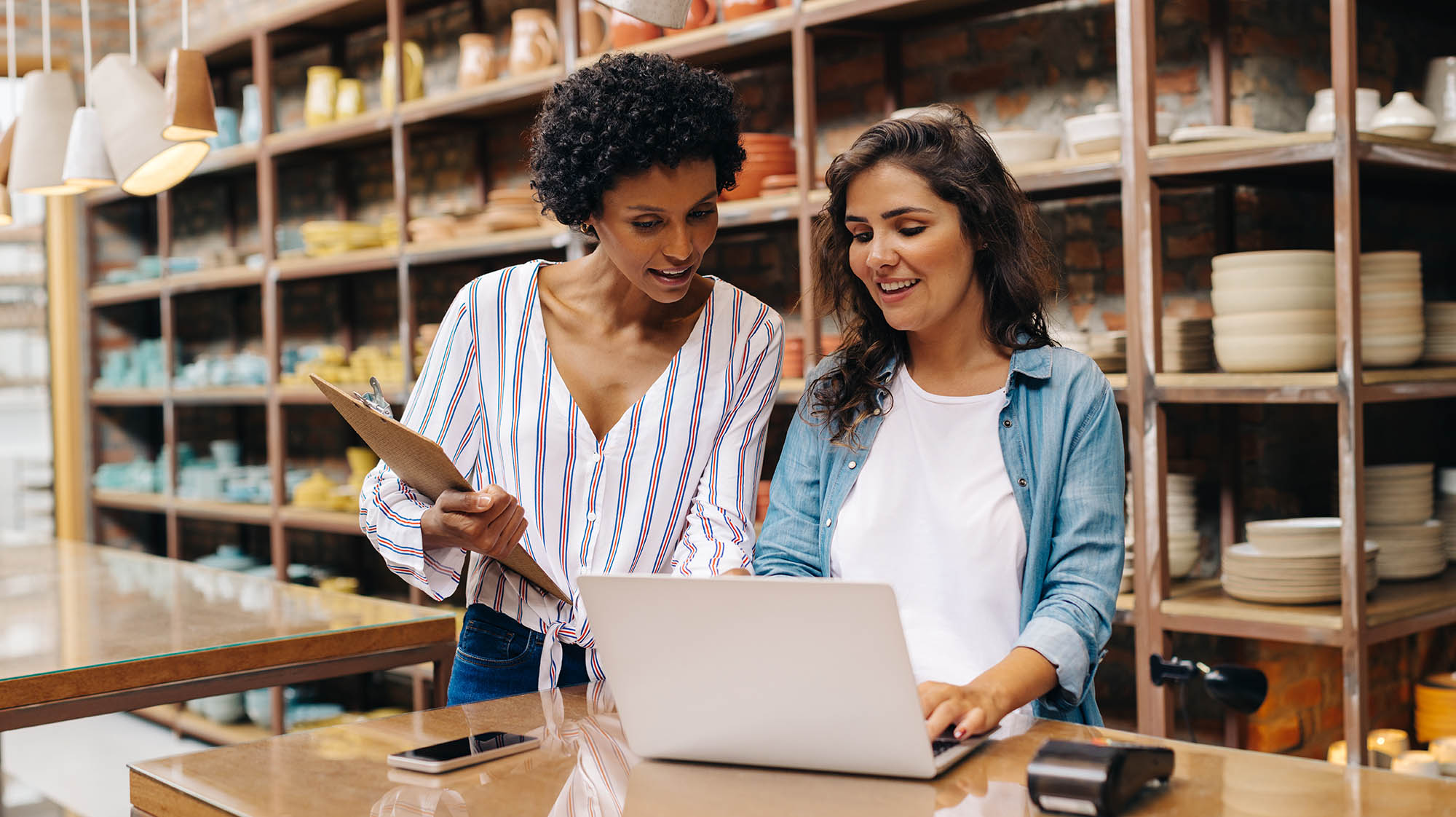 Two people collaborate in a retail setting, reviewing information on a laptop while standing behind a sales counter. Shelves of merchandise line the background, highlighting a small business environment.