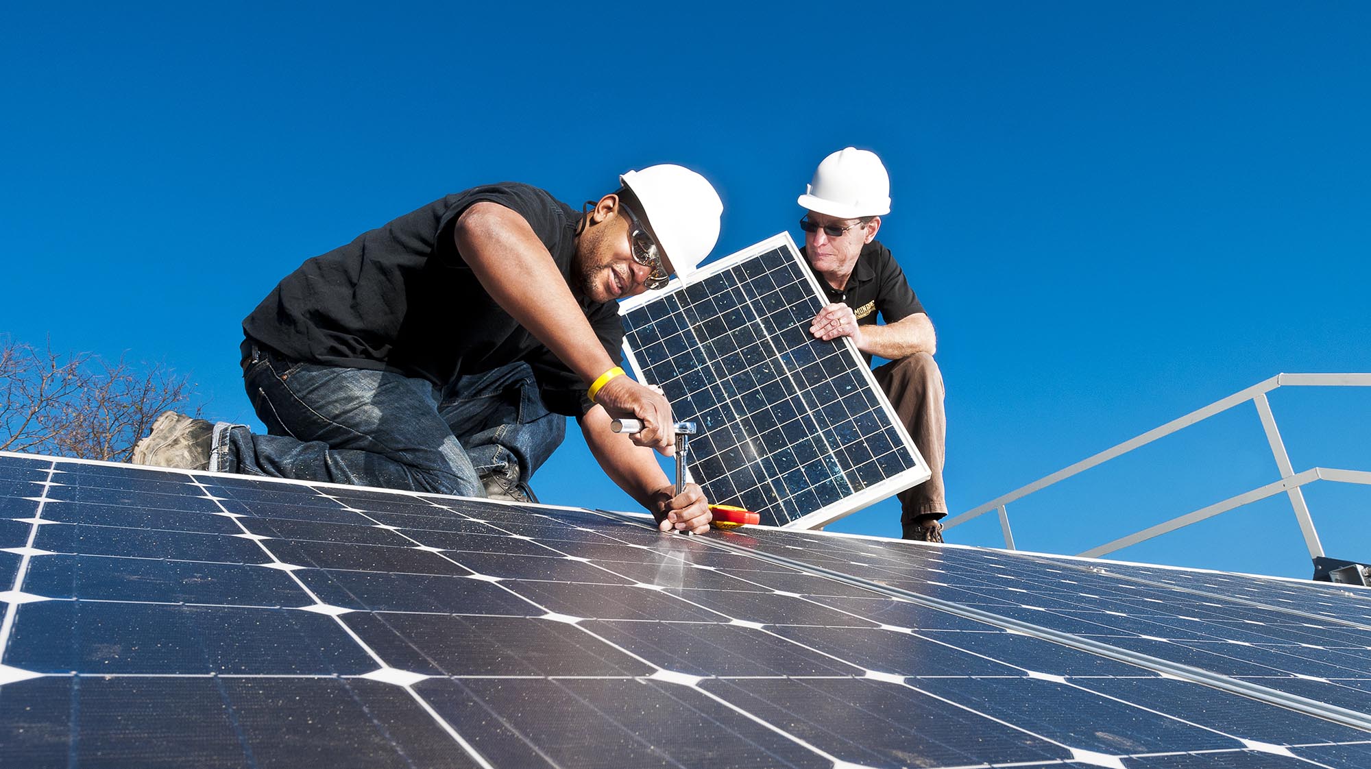 Two people wearing hard hats install and secure solar panels on a rooftop, demonstrating hands-on training in solar energy technology under a clear blue sky.