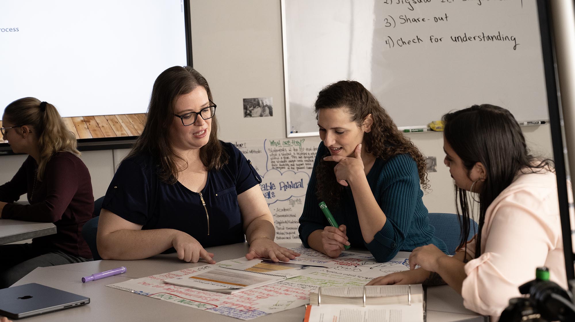 A teacher leans on a desk in a classroom
