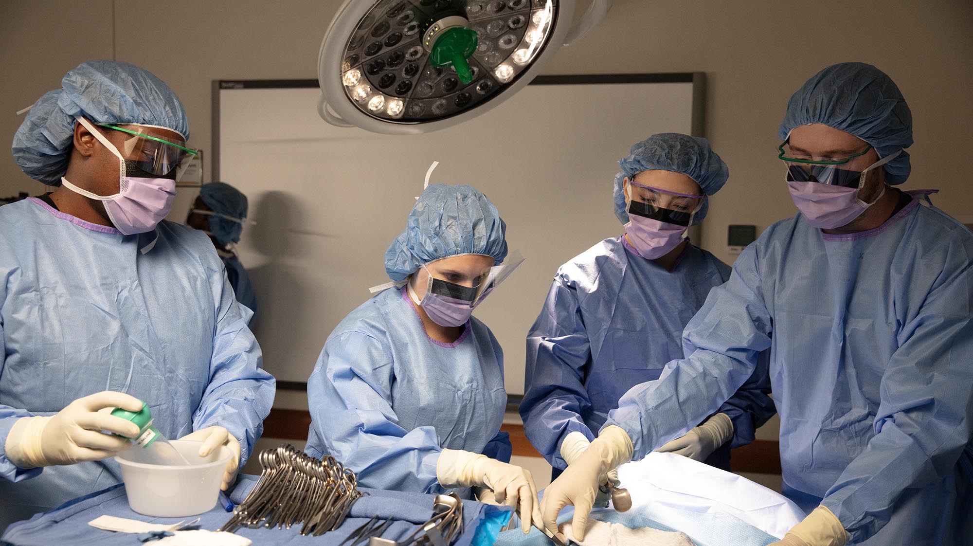 Surgical technology students wearing gowns, masks, gloves, and protective eyewear practice sterile surgical procedures together in a simulated operating room under surgical lights.