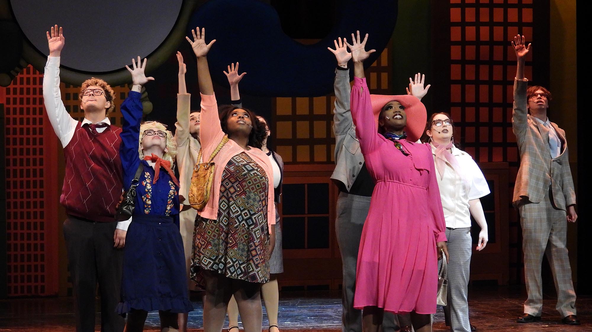 Students perform on stage during a theater production, wearing colorful costumes and raising their hands as part of a musical ensemble performance on a city-themed set.