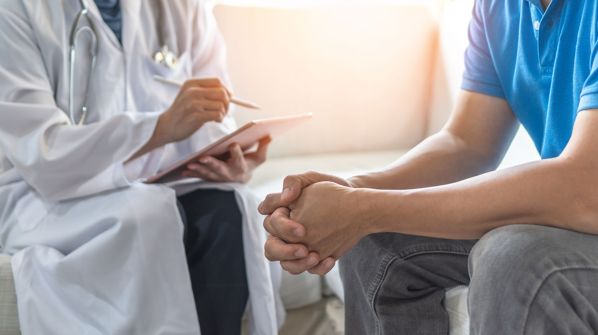 Healthcare professional consulting with a patient during an appointment, using a tablet to take notes while the patient sits with hands clasped in a supportive clinical setting.