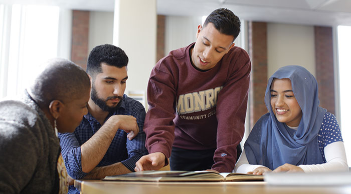 Three male students and one female student who is wearing a head scarf sitting at a table looking at a book