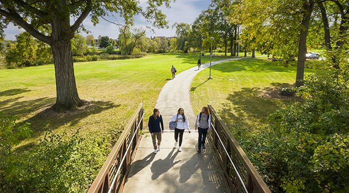 Three students walking outside on a wooden bridge with trees lining a walking path