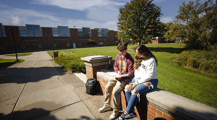 One male and one female student sitting outside on a bench looking at a textbook