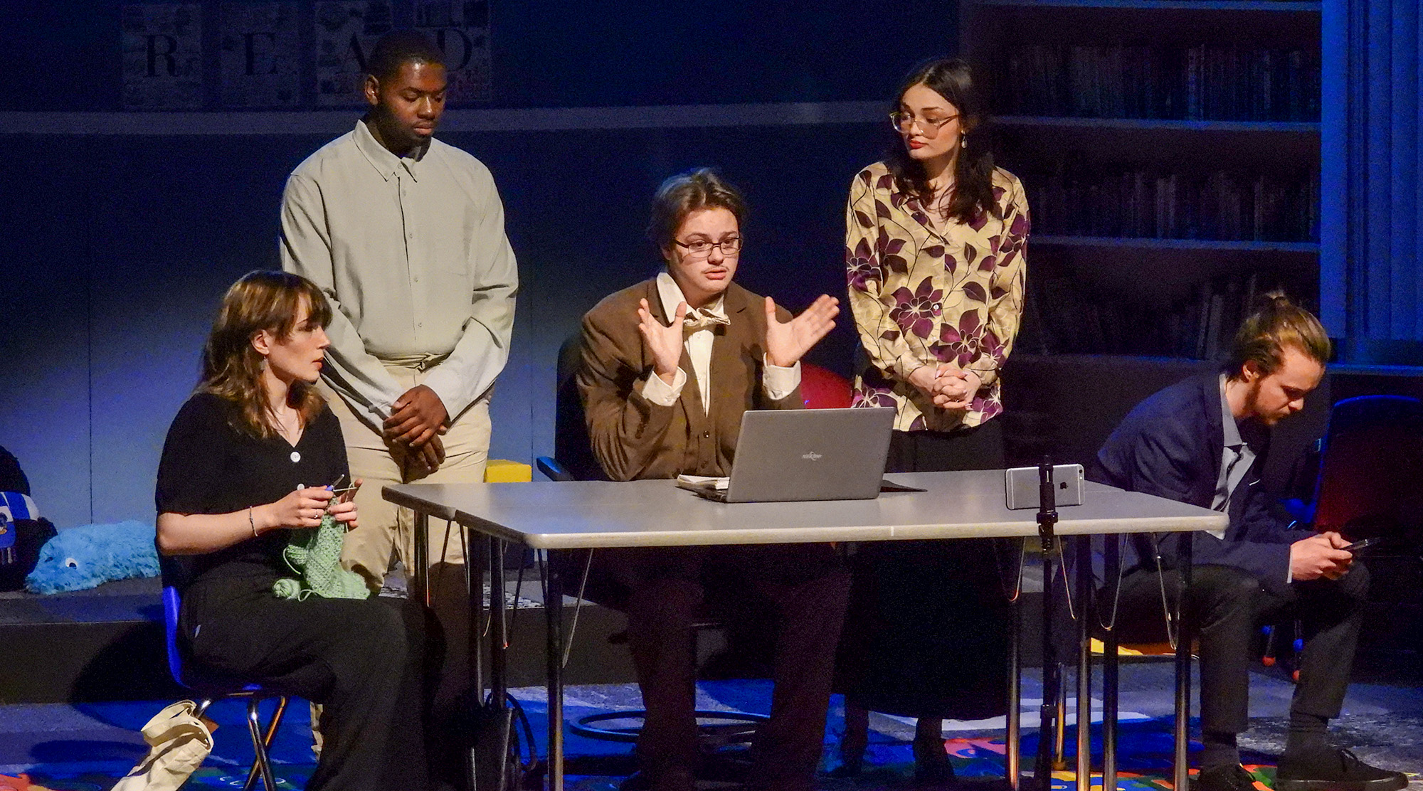 Five performers gather around a table on a dimly lit stage, with one speaking beside a laptop, while others knit, stand, or sit nearby under blue and purple lighting.