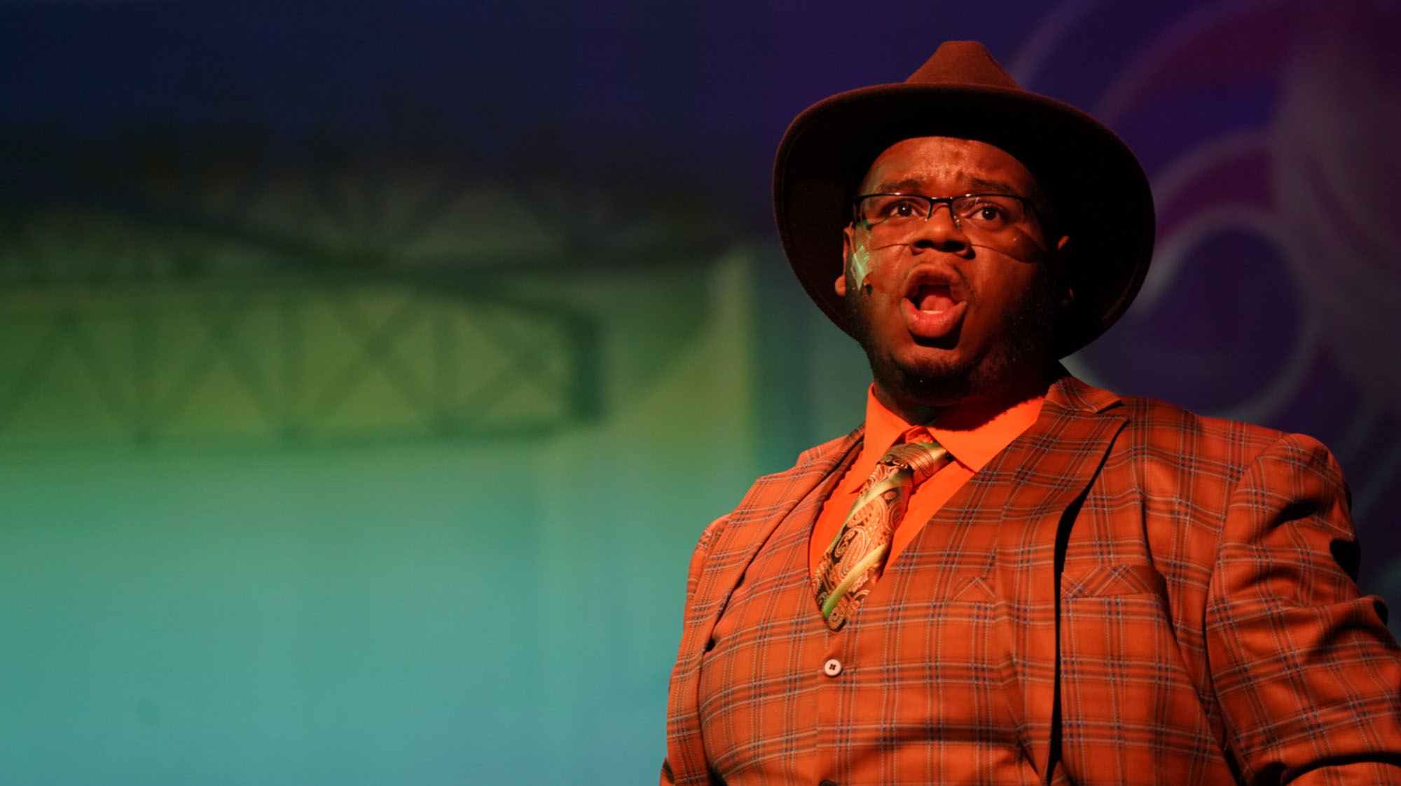 A performer in a colorful plaid suit, hat, and tie sings onstage under dramatic theatrical lighting.