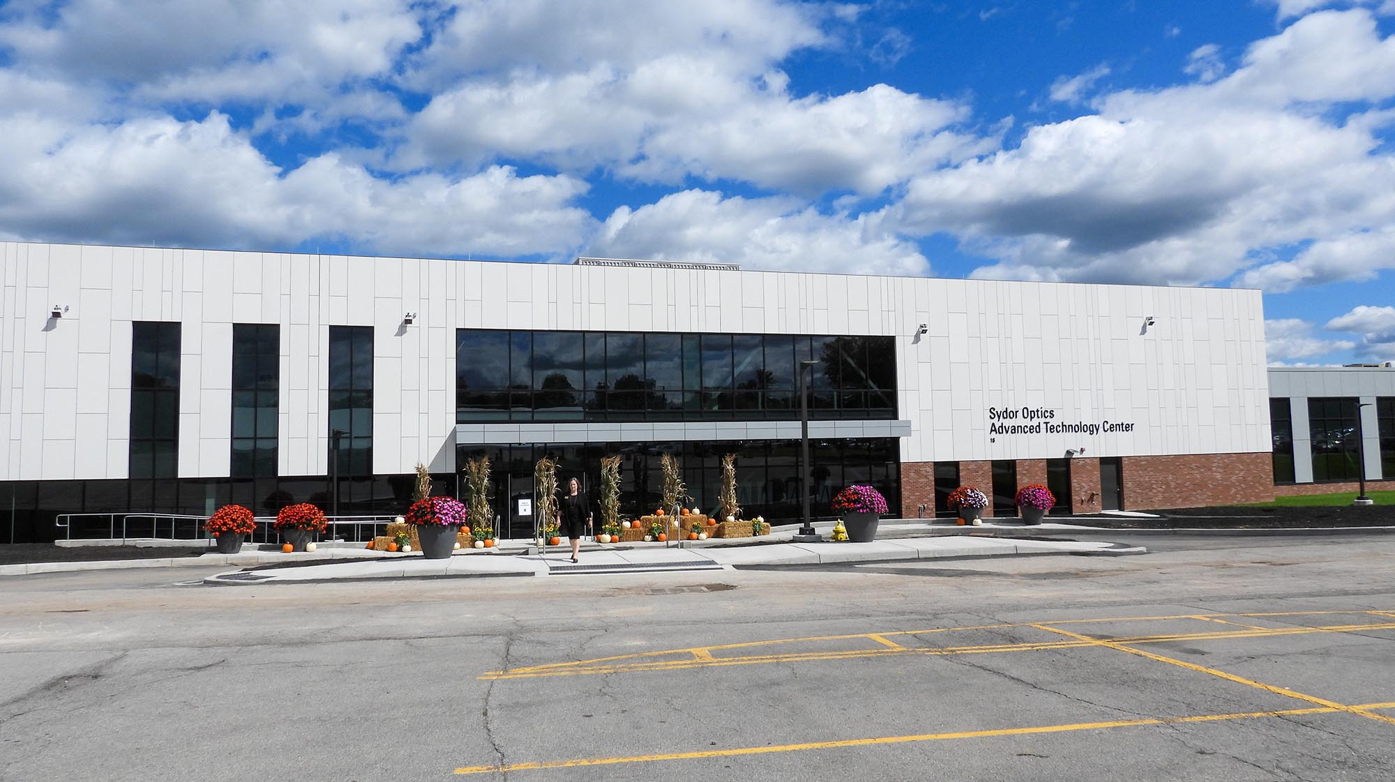 Exterior rendering of MCC's Sydor Optics Advanced Technology Center featuring a modern building with large windows, landscaped grounds, and outdoor seating.