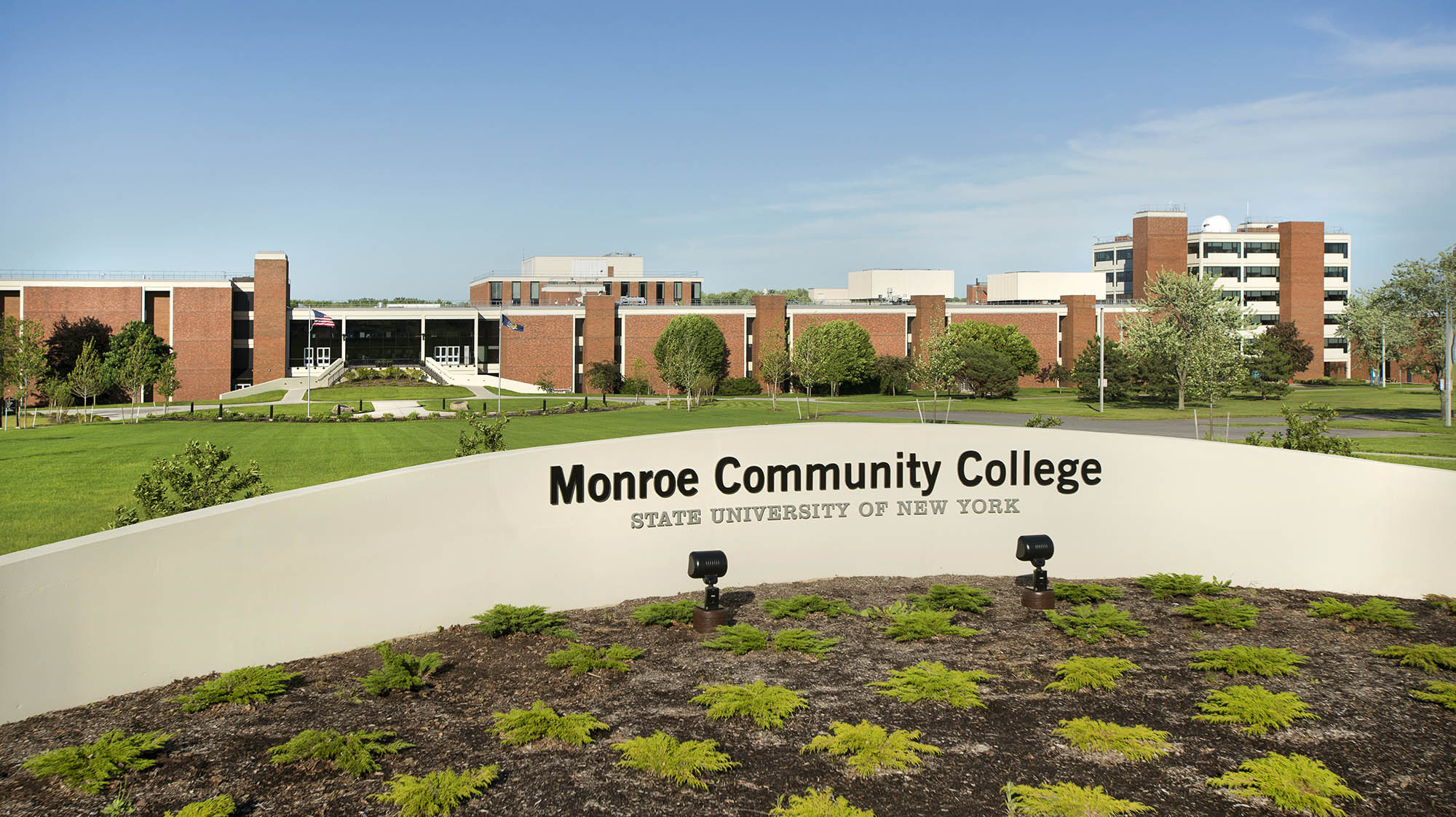 Monroe Community College campus with brick academic buildings, landscaped grounds, and a campus entrance sign.