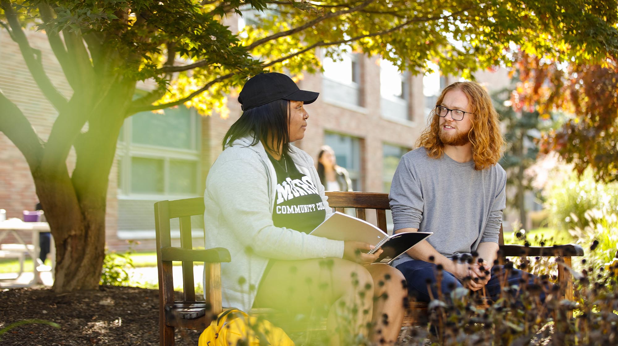 2 students sitting outside on a bench surrounded by fall foliage
