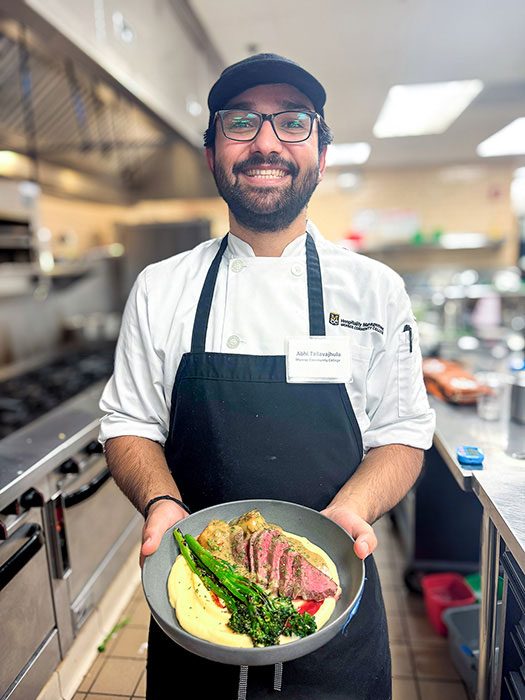 A smiling Monroe Community College hospitality management student chef presents a plated dish in a commercial kitchen.