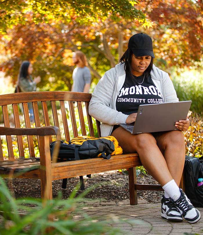 A Monroe Community College student studies on a laptop while sitting on a campus bench surrounded by trees in autumn.