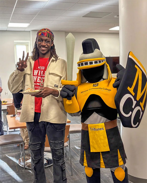 A Monroe Community College student poses with Tiberius, the college mascot, while holding a plate of food.