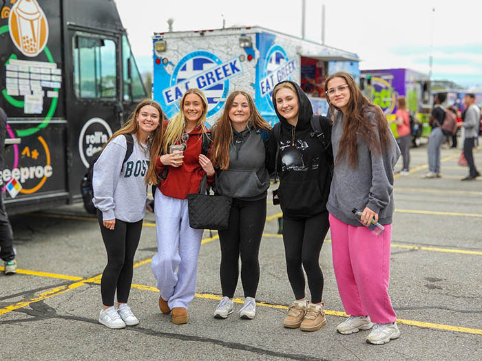 Five students pose together outdoors at a campus event with food trucks in the background.