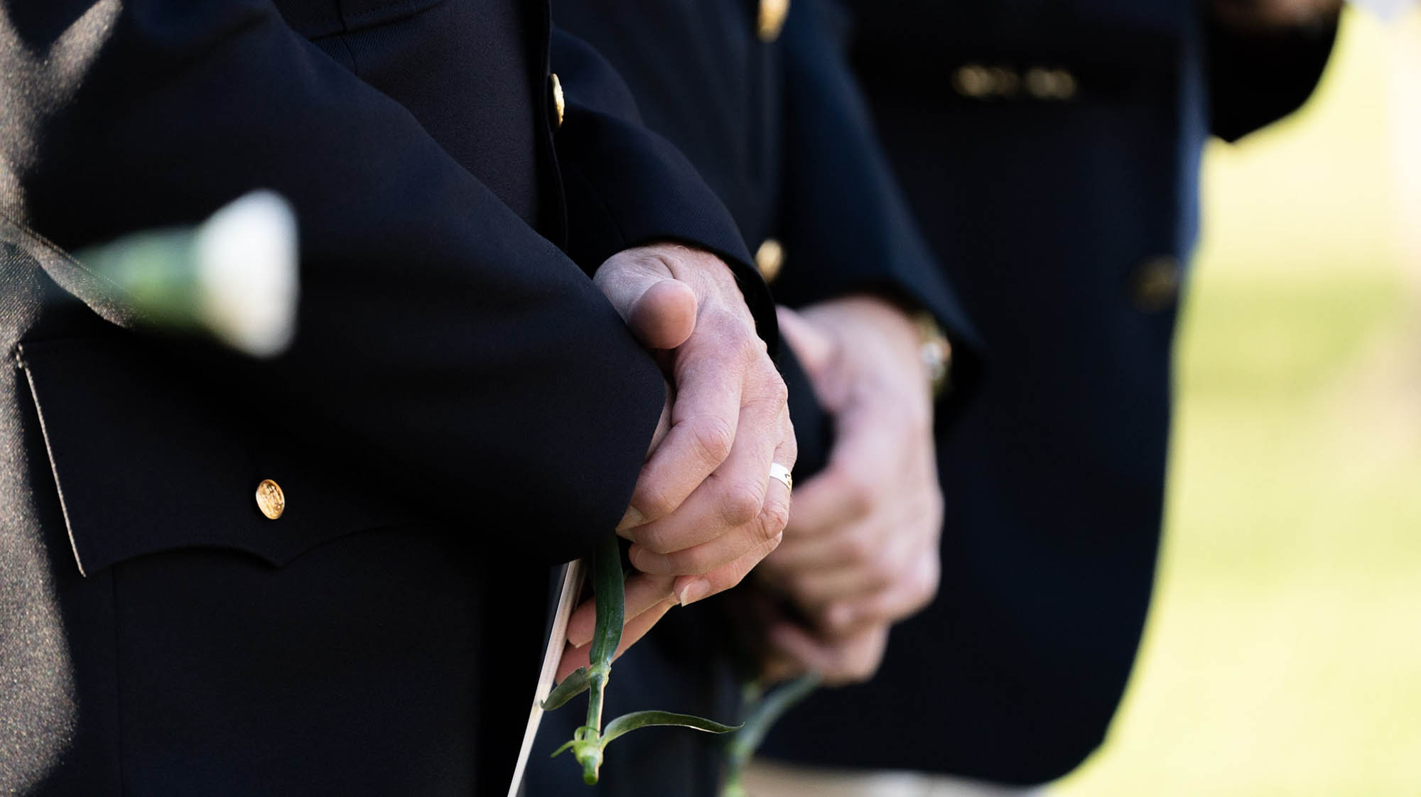 Close-up of a person in a dark uniform holding a green stem and white flower, with another person in a similar uniform in the background.