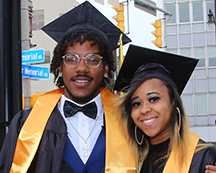 Male and female MCC students in cap and gown posing outside of the Blue Cross Arena after commencement exercises in Rochester, NY