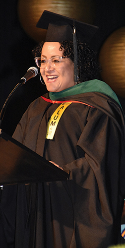 MCC Alumni Angelica Perez-Delgado, in cap and gown with green hood and “Alum” ribbon, speaking at 2024 MCC Commencement