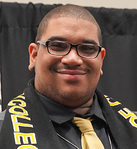 Portrait of MCC graduate Casan Curry standing in front of a black cloth backdrop and wearing a gray blazer and a scarf with the MCC logo over a black shirt and gold tie