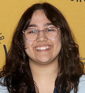 Portrait of MCC graduating student Elle Parrotta standing in front of a gold backdrop wearing a light blue top and a blue ribbon around her neck