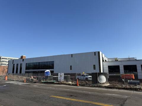 A large, modern building under construction with a white exterior is seen behind a chain-link fence. The area in front of the building is fenced off with construction equipment. There's a clear blue sky overhead.