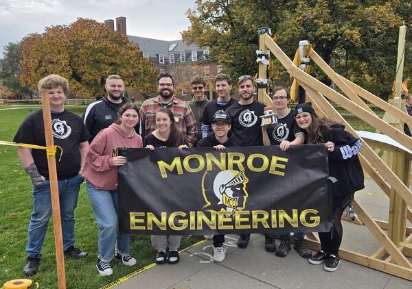 MCC's Engineering Club students holding a black Monroe Banners while standing next to a 2x4 wood structure.