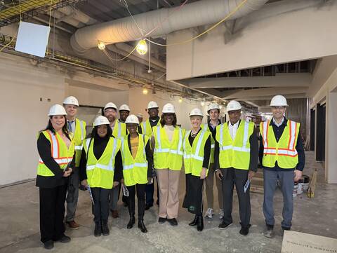 A group of individuals wearing hard hats and high-visibility vests stands in a construction area. The environment includes visible wiring, lighting, and unfinished surfaces. They're posing for a group photo.