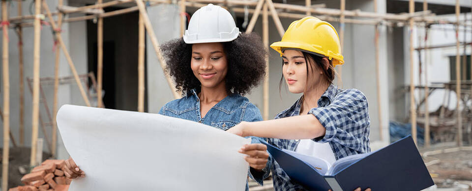 Two women wearing hardhats reviewing blueprints on a construction site